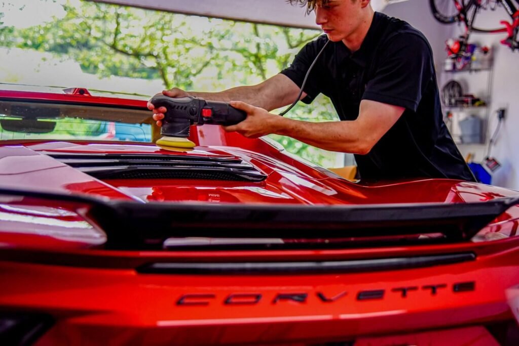 Guy with a dual action polisher on the back roof of red corvette in a garage